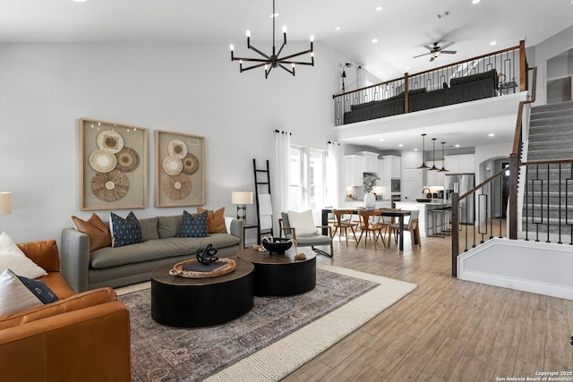 living area featuring stairs, light wood finished floors, visible vents, a high ceiling, and ceiling fan with notable chandelier