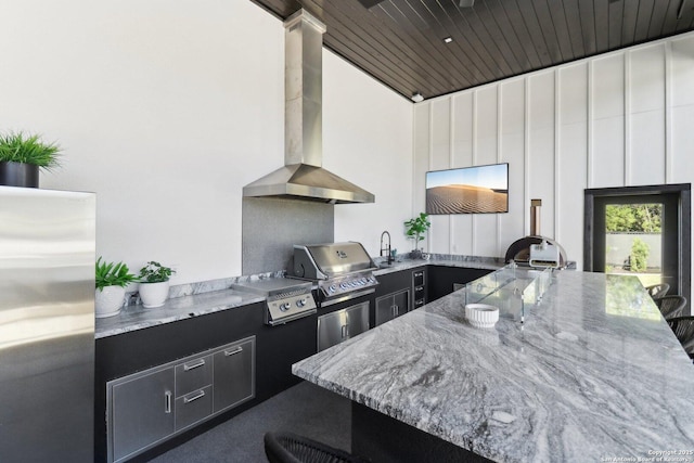 kitchen with freestanding refrigerator, a sink, light stone countertops, dark cabinets, and wall chimney exhaust hood