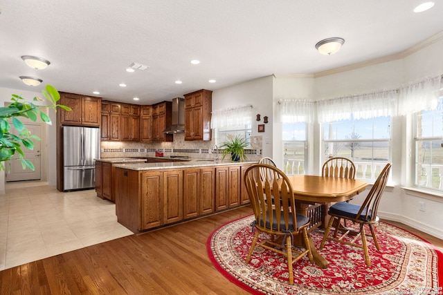 kitchen with light wood-style floors, freestanding refrigerator, a peninsula, wall chimney range hood, and backsplash