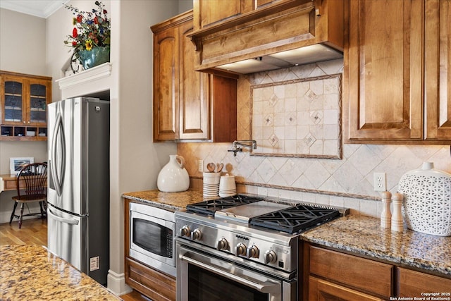 kitchen featuring appliances with stainless steel finishes, custom exhaust hood, light stone countertops, brown cabinetry, and crown molding