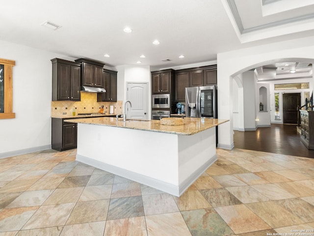 kitchen featuring arched walkways, stainless steel appliances, dark brown cabinets, under cabinet range hood, and backsplash