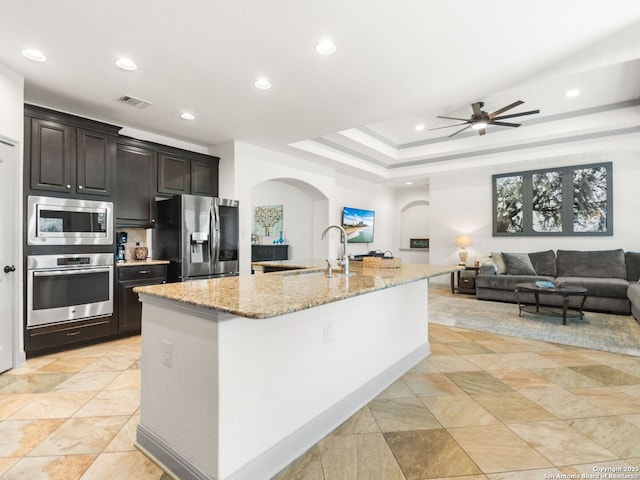 kitchen with a center island with sink, a sink, light stone counters, stainless steel appliances, and arched walkways