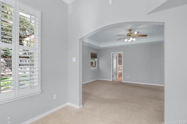 carpeted empty room with arched walkways, a raised ceiling, a ceiling fan, and baseboards