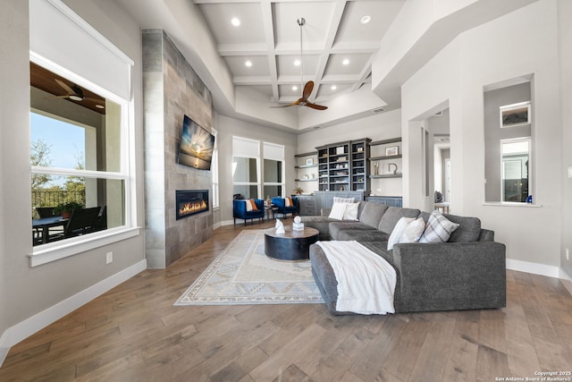 living area with a tiled fireplace, ceiling fan, wood finished floors, coffered ceiling, and baseboards