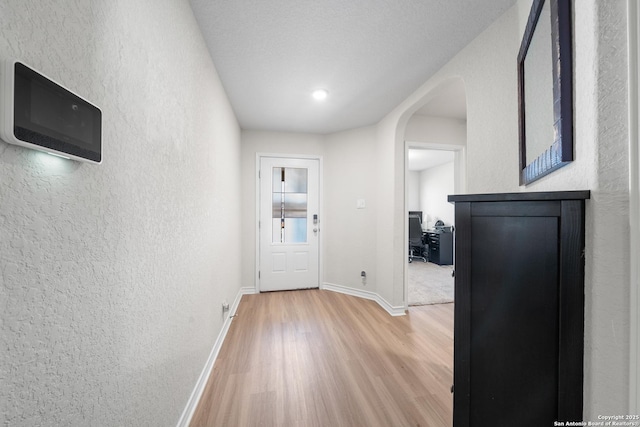 corridor featuring arched walkways, a textured wall, baseboards, and light wood-style floors