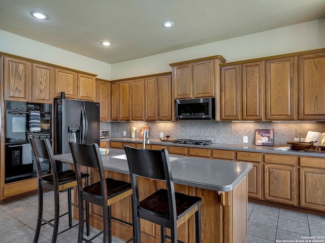 kitchen featuring a center island with sink, dark countertops, brown cabinets, black appliances, and backsplash