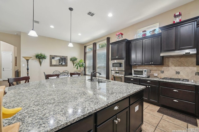 kitchen with light stone counters, under cabinet range hood, stainless steel appliances, a sink, and backsplash