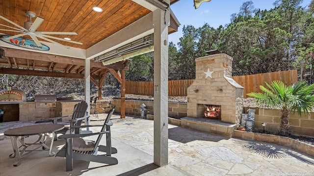 view of patio / terrace featuring an outdoor stone fireplace, area for grilling, fence, a ceiling fan, and grilling area