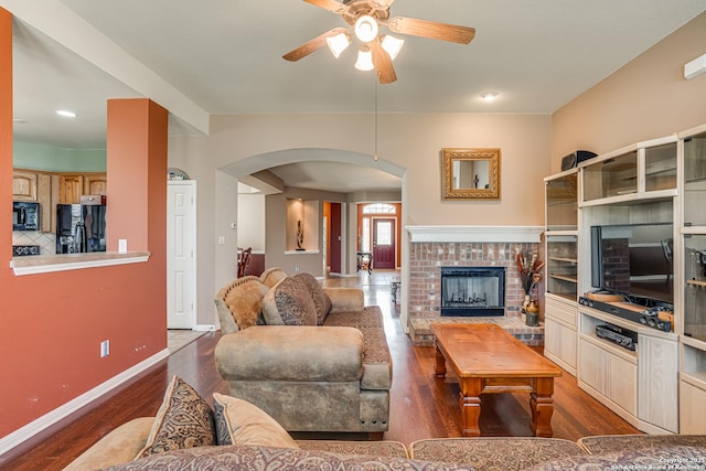 living area featuring arched walkways, a ceiling fan, baseboards, a brick fireplace, and dark wood finished floors