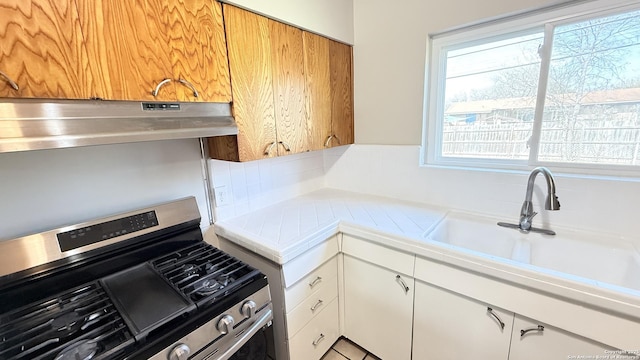 kitchen with white cabinets, tile countertops, under cabinet range hood, a sink, and gas stove