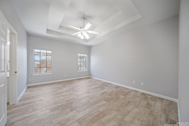 empty room with light wood-type flooring, a tray ceiling, baseboards, and a ceiling fan