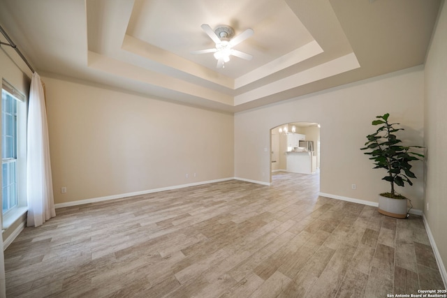 empty room featuring ceiling fan, baseboards, a tray ceiling, light wood-style floors, and arched walkways