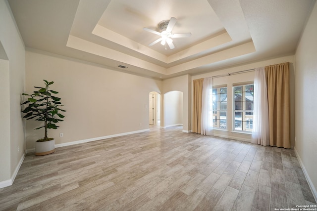 spare room with baseboards, a tray ceiling, arched walkways, ceiling fan, and light wood-type flooring