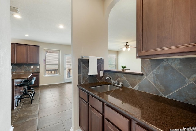 kitchen with light tile patterned floors, arched walkways, a sink, backsplash, and dark stone counters