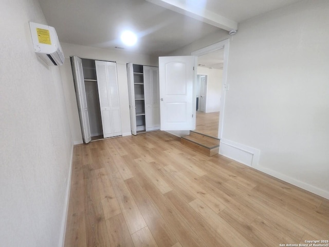 unfurnished bedroom with light wood finished floors, two closets, a wall mounted AC, beamed ceiling, and baseboards