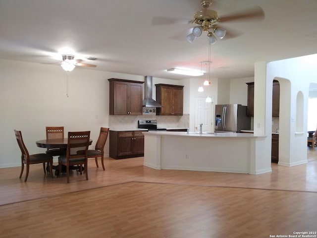 kitchen featuring arched walkways, light wood-style floors, stainless steel appliances, wall chimney range hood, and backsplash