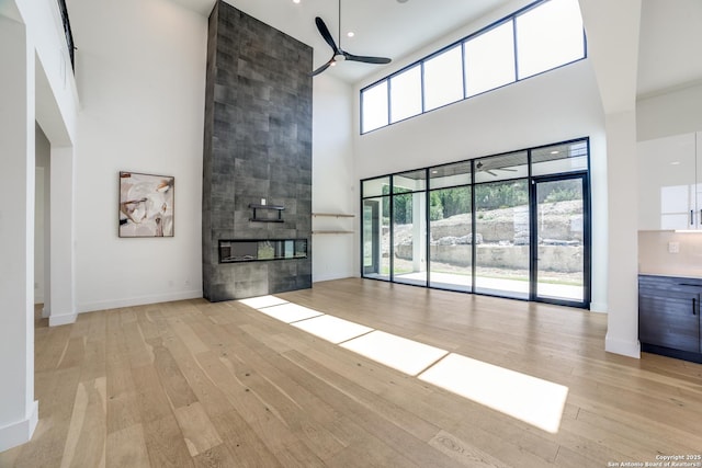 unfurnished living room with ceiling fan, a fireplace, a towering ceiling, baseboards, and light wood-style floors