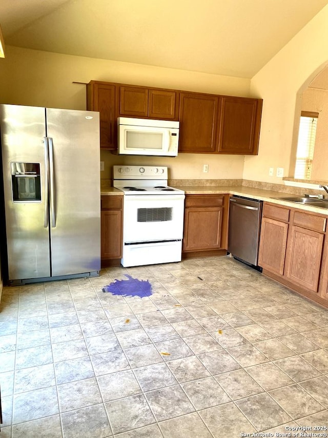 kitchen featuring arched walkways, a sink, vaulted ceiling, appliances with stainless steel finishes, and brown cabinetry