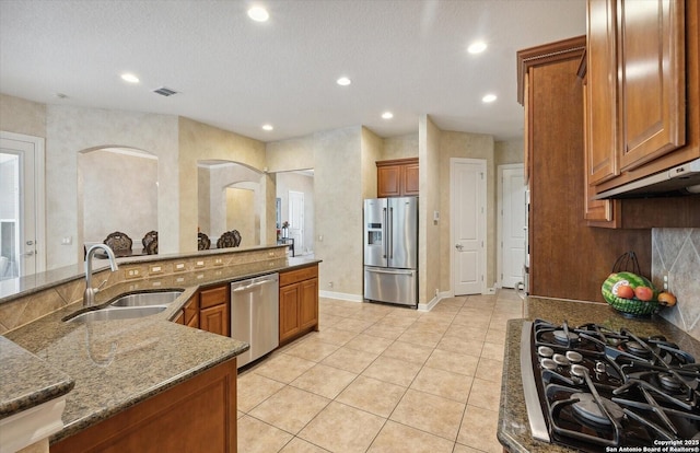 kitchen with light tile patterned flooring, a sink, appliances with stainless steel finishes, brown cabinets, and dark stone counters