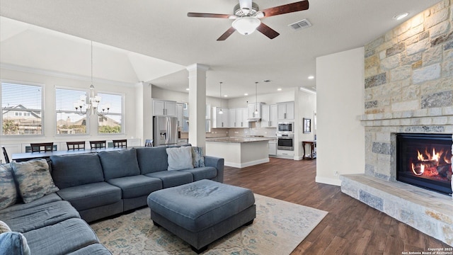 living area featuring visible vents, a stone fireplace, ceiling fan with notable chandelier, ornate columns, and dark wood-style flooring