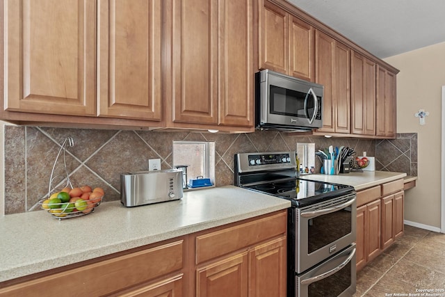 kitchen with baseboards, light countertops, appliances with stainless steel finishes, brown cabinets, and backsplash