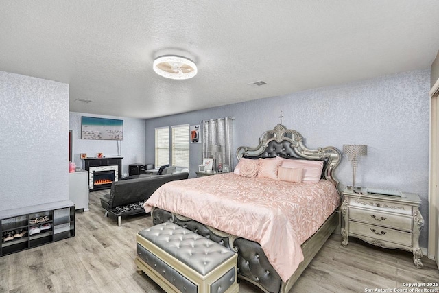 bedroom with wood finished floors, visible vents, a textured wall, and a lit fireplace