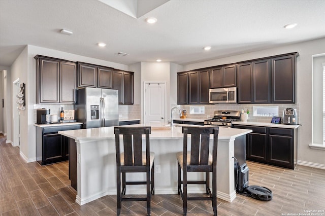 kitchen with dark brown cabinets, wood finish floors, an island with sink, light countertops, and appliances with stainless steel finishes