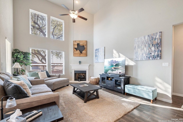 living area with a fireplace with raised hearth, a ceiling fan, wood finished floors, baseboards, and a towering ceiling