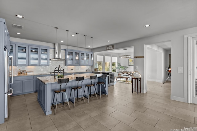 kitchen with light stone counters, visible vents, a kitchen island with sink, wall chimney exhaust hood, and tasteful backsplash