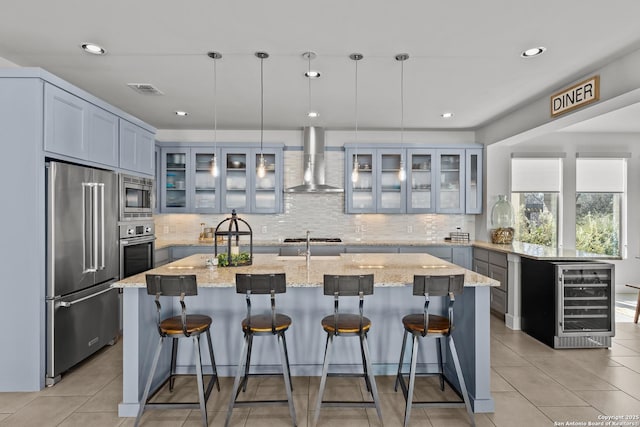 kitchen featuring light tile patterned floors, a center island with sink, wine cooler, appliances with stainless steel finishes, and wall chimney exhaust hood