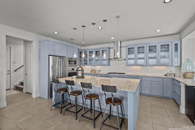 kitchen with a center island with sink, stainless steel appliances, wall chimney range hood, decorative backsplash, and light stone countertops