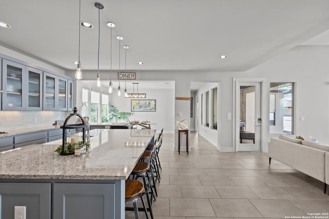 kitchen with gray cabinetry, a kitchen breakfast bar, tasteful backsplash, glass insert cabinets, and light stone countertops