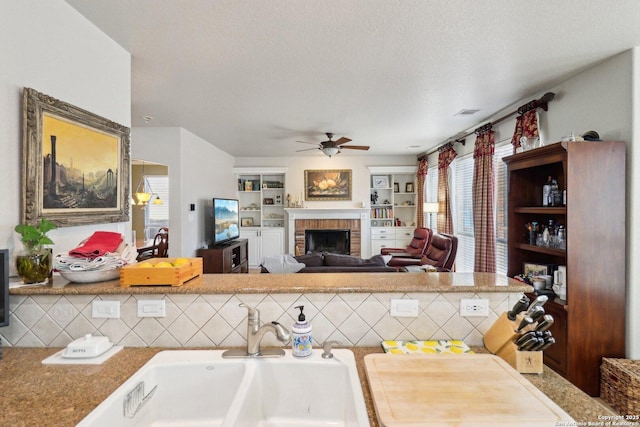 kitchen featuring backsplash, a brick fireplace, ceiling fan, a textured ceiling, and a sink