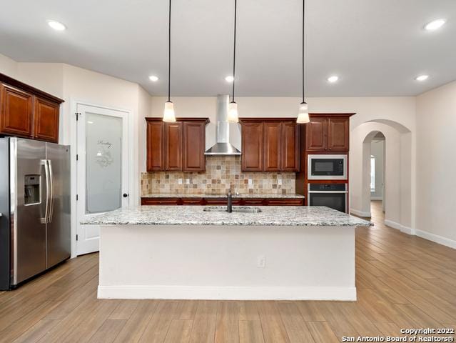 kitchen with a sink, light stone counters, arched walkways, appliances with stainless steel finishes, and wall chimney exhaust hood