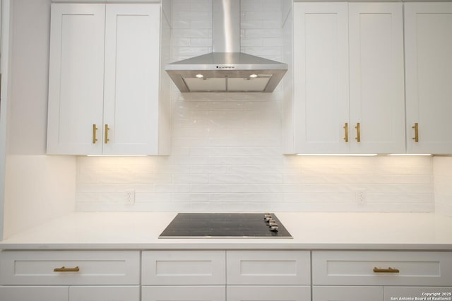 kitchen featuring black electric stovetop, light countertops, white cabinets, wall chimney range hood, and backsplash