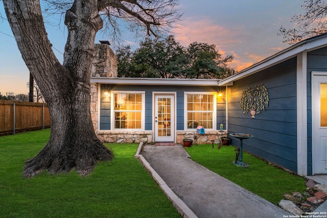 exterior entry at dusk with stone siding, a lawn, a chimney, and fence