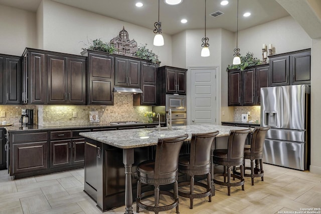 kitchen with visible vents, a center island with sink, light stone counters, stainless steel appliances, and a high ceiling