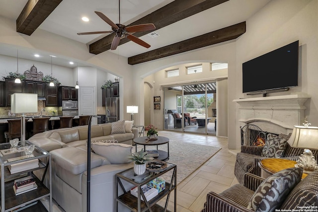 living area featuring beamed ceiling, visible vents, a warm lit fireplace, arched walkways, and ceiling fan