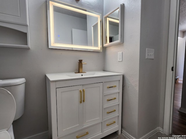 bathroom with vanity, toilet, a textured wall, and baseboards