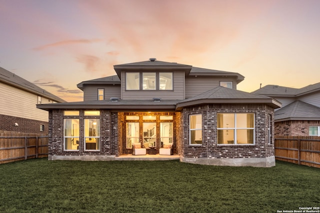 back of property at dusk featuring brick siding, a fenced backyard, a lawn, and a patio area