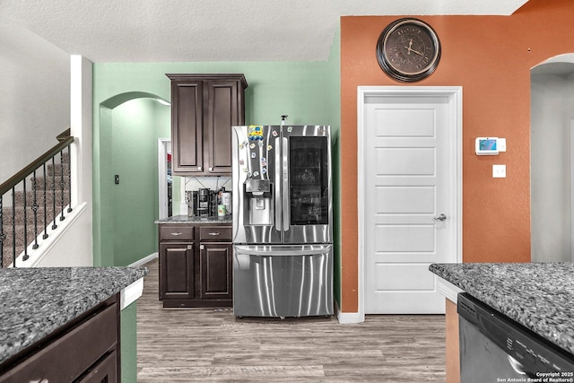 kitchen with dark stone countertops, stainless steel appliances, arched walkways, dark brown cabinetry, and light wood finished floors