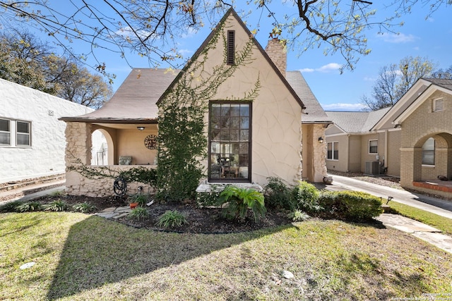 tudor-style house featuring stucco siding, a chimney, central AC, and a front lawn