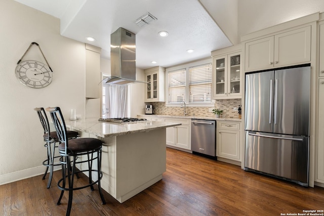 kitchen featuring a peninsula, island exhaust hood, stainless steel appliances, dark wood-type flooring, and a kitchen bar