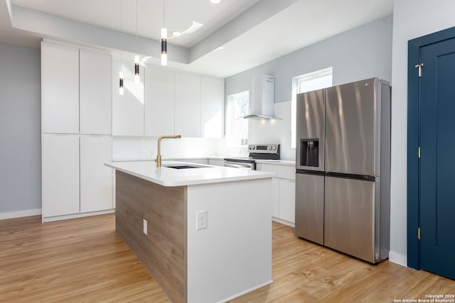 kitchen with wall chimney range hood, light wood-style flooring, appliances with stainless steel finishes, white cabinetry, and a sink