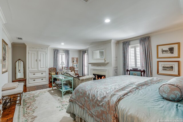 bedroom with dark wood-type flooring, recessed lighting, a warm lit fireplace, and ornamental molding