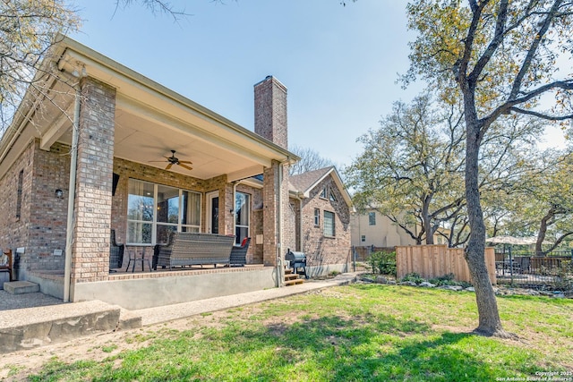 rear view of house with a lawn, a ceiling fan, fence, brick siding, and a chimney