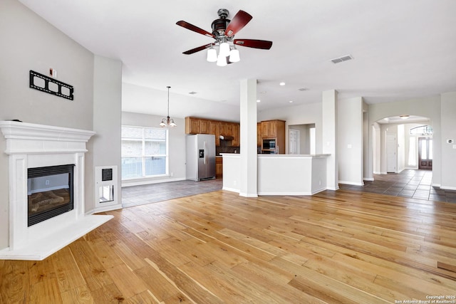 unfurnished living room featuring visible vents, ceiling fan with notable chandelier, a glass covered fireplace, arched walkways, and light wood finished floors