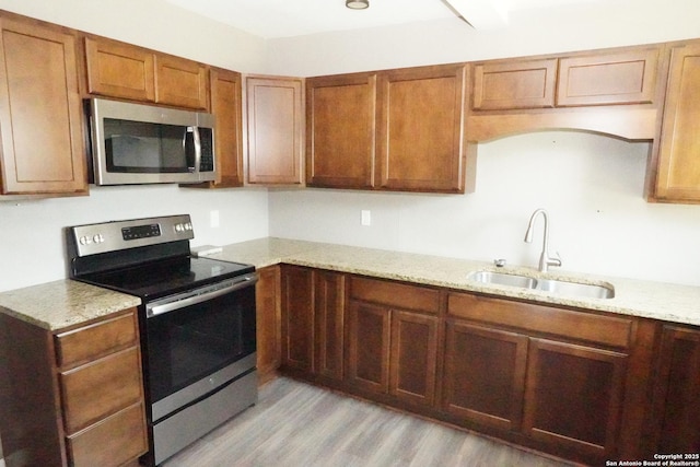 kitchen featuring light wood finished floors, light stone counters, brown cabinetry, stainless steel appliances, and a sink