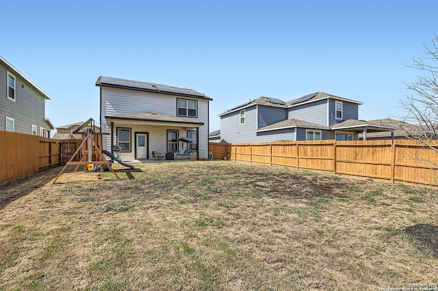 rear view of property with a lawn, roof mounted solar panels, a fenced backyard, a playground, and a patio area