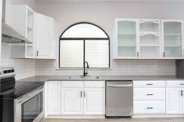 kitchen featuring glass insert cabinets, wall chimney range hood, appliances with stainless steel finishes, white cabinetry, and a sink